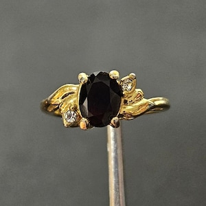 May include: A gold-toned ring featuring a central oval-cut dark gemstone flanked by two small, clear, round-cut accent stones. The band has a polished finish with a decorative, flowing metal design surrounding the central setting.