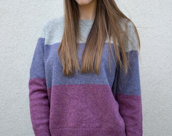 May include: A woman wearing a striped sweater with grey, purple, and pink horizontal stripes. The sweater has long sleeves and a relaxed fit.