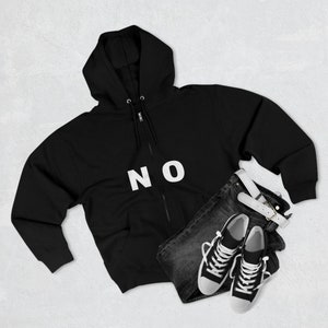 Puede incluir: Sudadera negra con cremallera y capucha con la palabra "NO" en blanco impresa en la parte delantera. La sudadera se combina con unos vaqueros oscuros y zapatillas blancas.