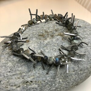 May include: A silver bracelet with a black leather cord and iridescent gray gemstones. The bracelet is made of small, irregular pieces of leather that are connected by silver rings. The gemstones are scattered throughout the bracelet, creating a unique and eye-catching design.