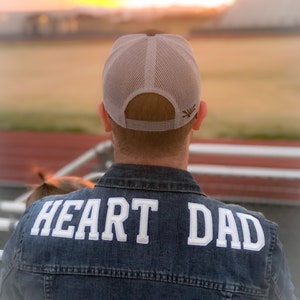 以下が含まれることがあります： 青いデニムジャケットを着て、背中に「HEART DAD」という白い文字が書かれた人。白いメッシュの野球帽をかぶり、茶色のロゴが付いている。