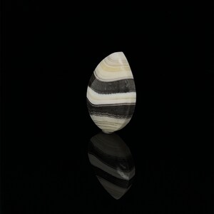 Puede incluir: Un cabuj&oacute;n de &aacute;gata con bandas blancas y negras en forma de l&aacute;grima. La piedra est&aacute; pulida y tiene una superficie lisa.
