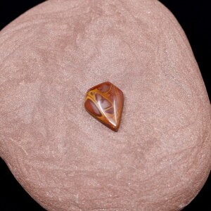 May include: A brown and yellow agate cabochon with a diamond shape. The cabochon is sitting on a brown stone.