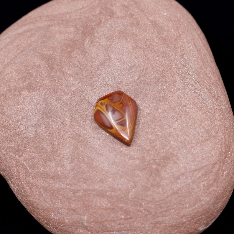 May include: A brown and yellow agate cabochon with a diamond shape. The cabochon is sitting on a brown stone.