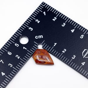 May include: A red, brown, and white agate cabochon with a pointed shape, measuring approximately 1.5 inches in length. The cabochon is sitting on a black ruler.