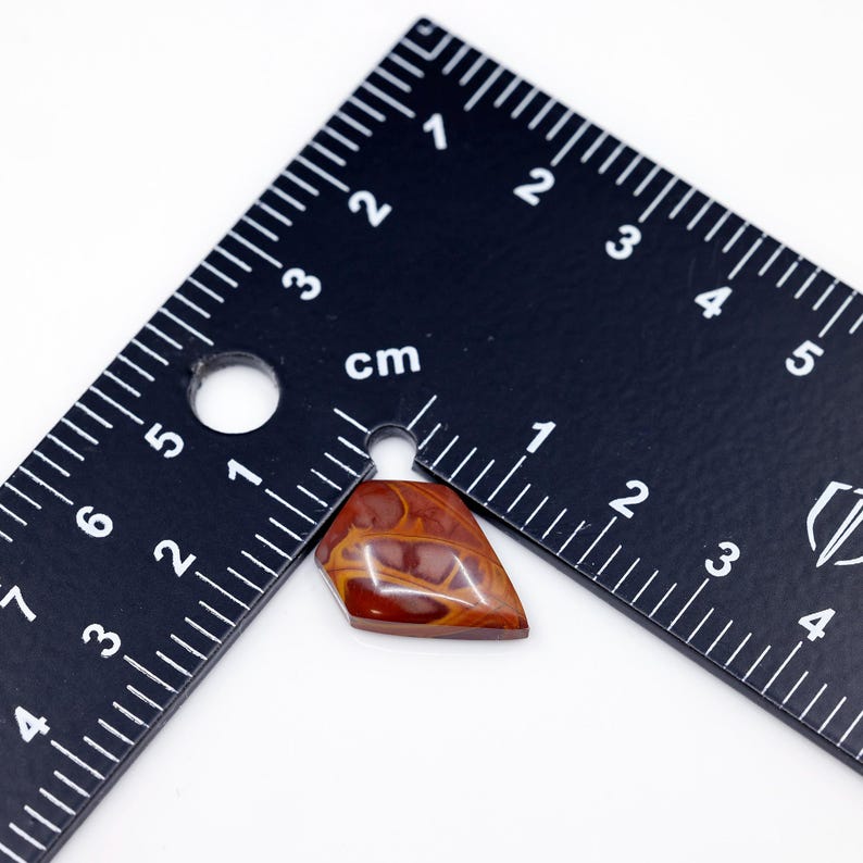 May include: A red, brown, and white agate cabochon with a pointed shape, measuring approximately 1.5 inches in length. The cabochon is sitting on a black ruler.