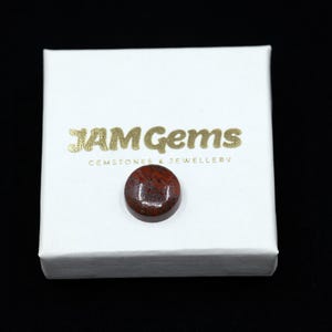 May include: A round, polished gemstone with a mottled red and brown pattern rests on a white box. The box has gold text that reads "JAM Gems GEMSTONES & JEWELLERY". The gemstone is the focal point, set against a dark background.