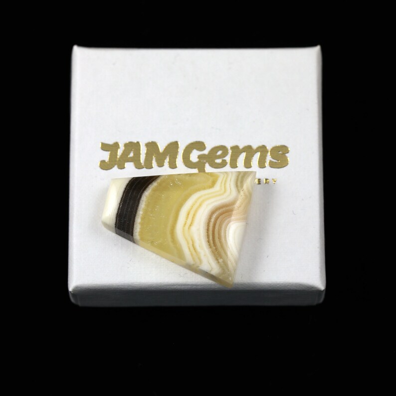 May include: A yellow, white, and black banded agate cabochon in a white box with the text "JAM Gems" printed in gold.