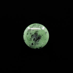 May include: A round, polished gemstone with a speckled green surface and dark black inclusions. The stone is set against a black background, highlighting its natural colors and textures. This gemstone is a decorative item.