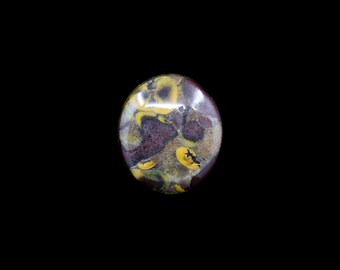 May include: Oval-shaped gemstone with a polished surface. The stone features a mix of colors, including yellow, brown, and purple, creating a unique pattern. The gemstone is set against a black background, highlighting its natural beauty.