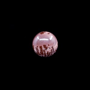May include: A polished, spherical gemstone with a mix of colors. The orb features a base of light pink with swirling patterns and patches of dark brown. The gemstone is set against a black background.