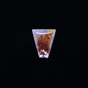 May include: A polished gemstone with a unique, abstract pattern. The stone features a triangular shape with a translucent, light purple base. Within the stone, there are intricate patterns of brown, orange, and white, creating a natural, artistic design.