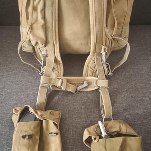 May include: Tan canvas backpack with multiple compartments and adjustable straps. Includes two side pouches and a lower pouch, all in matching tan. The backpack has a vintage military style.