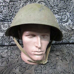 Puede incluir: Un casco militar verde oliva con una corona redondeada y un borde. El casco tiene una correa para la barbilla y se muestra en una cabeza de maniquí. El fondo presenta un patrón floral en blanco y negro.