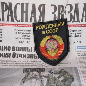 Puede incluir: Un parche negro con el texto "РОЖДЕННЫЙ В СССР" (Nacido en la URSS) y un emblema soviético, sobre un fondo de periódico. El parche presenta una corona dorada y un emblema rojo y dorado.