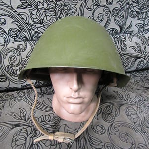 May include: An olive-green military helmet with a rounded top and a leather chin strap. The helmet is displayed on a mannequin head. The background features a black and white floral pattern.