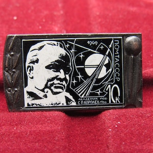 Puede incluir: Una taza de cerámica blanca con asa negra y el texto "But First, Coffee" en negrita negra. La taza se encuentra sobre un fondo borroso, sugiriendo un ambiente acogedor.