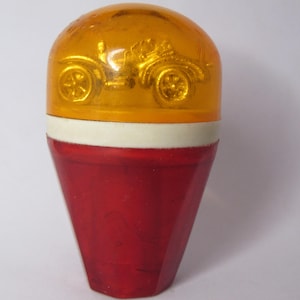 May include: A vintage bottle stopper with a red, cone-shaped base and a yellow top featuring a car design. A white band separates the two sections. The stopper is likely made of glass or a similar material.