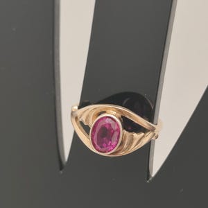 May include: A gold ring featuring a central oval-shaped, vibrant pink gemstone set in a smooth bezel. The band has a split design that curves around the stone, creating an eye-like shape. The metal has a warm, polished finish throughout the piece.