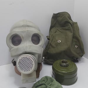 Könnte beinhalten: Ein Vintage-Gasmasken-Set mit einer hellgrauen Maske mit großen Augenöffnungen und einem kreisförmigen Filter. Das Set enthält eine grüne Canvas-Tasche, einen grünen Filterkanister und weiteres Zubehör. Die Artikel werden auf einer weißen Oberfläche präsentiert.