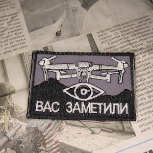 Puede incluir: Parche bordado con un diseño de dron sobre un ojo, con el texto ruso "ВАС ЗАМЕТИЛИ" (Te han notado). El parche es negro, gris y blanco, con forma rectangular.