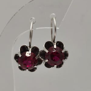May include: A pair of silver-toned hoop earrings, each featuring a flower-shaped design. The flower petals are black, surrounding a central, round, deep red gemstone. The earrings are displayed against a neutral background.