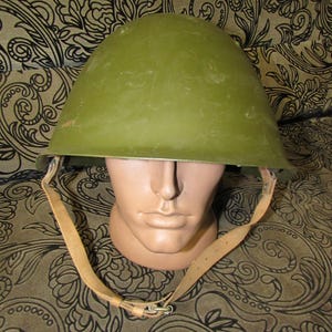 May include: An olive green military helmet with a leather chin strap. The helmet is displayed on a mannequin head. The helmet has a rounded shape and a slight sheen. The chin strap is a light tan color.