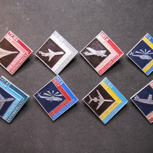 Pode incluir: Oito pinos de aviação vintage com designs em forma de diamante. Cada pino apresenta um avião ou helicóptero prateado contra um fundo preto, emoldurado por bordas coloridas em vermelho, azul, amarelo e branco. Texto em escrita cirílica visível.