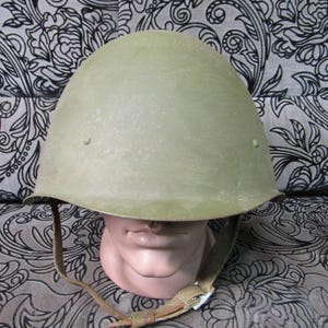 Puede incluir: Un casco militar verde oliva con barboquejo. El casco se exhibe sobre una cabeza de maniquí. El fondo presenta un estampado floral en blanco y negro.