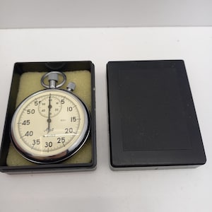 May include: A vintage stopwatch with a cream-colored face, black numerals, and a chrome bezel. The stopwatch is in a black case with a yellow interior. The case is open, revealing the stopwatch. The stopwatch has a button on top and a dial on the side.