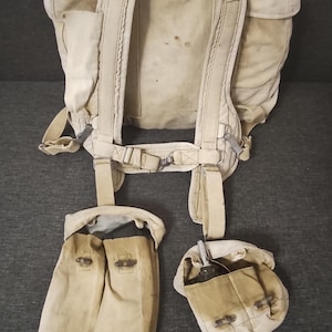 May include: A beige canvas military-style backpack featuring adjustable shoulder straps and a waist belt. Attached to the lower section are two separate canvas pouches, one elongated and one smaller, both secured with metal toggles and fasteners.