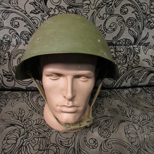 Puede incluir: Un casco militar verde oliva con una parte superior redondeada y un borde. El casco está asegurado con una correa de barbilla beige y se muestra en una cabeza de maniquí. El fondo presenta un patrón floral negro y gris.