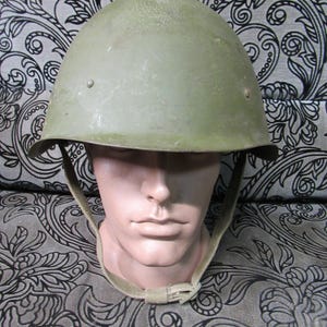 Puede incluir: Un casco militar verde oliva con la parte superior redondeada y un borde. El casco tiene una correa para la barbilla y se muestra en una cabeza de maniquí. La superficie del casco muestra signos de desgaste. El fondo es una tela estampada.