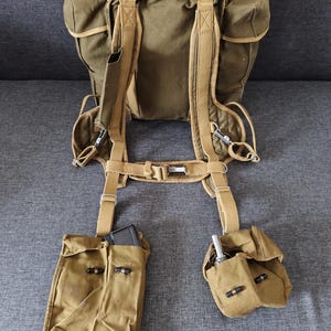 May include: Olive green canvas backpack with tan straps and two attached pouches. The backpack has a large main compartment and adjustable straps. The pouches are secured with black closures. A magazine and a small metal object are visible inside the pouches.