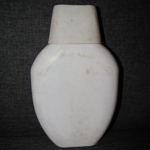 May include: A white, ceramic vase with a rounded body and a rectangular top. The vase has a smooth surface with some discoloration. The design is simple and modern, suitable for home decor.