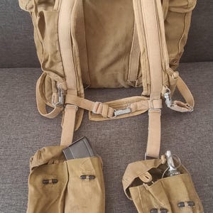 May include: A tan canvas military-style backpack featuring adjustable shoulder straps and a waist belt. Attached to the lower straps are two separate canvas pouches, one containing a dark rectangular magazine and the other a metal hand grenade.