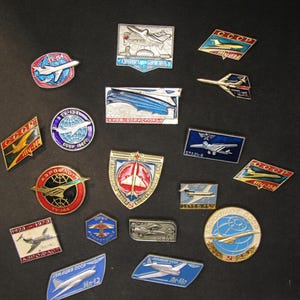 Pode incluir: Uma coleção de pinos de aviação da era soviética vintage. Os pinos apresentam vários designs de aeronaves, o logotipo CCCP e texto cirílico. Os pinos têm várias formas, incluindo círculos, quadrados e escudos, com uma gama de cores.