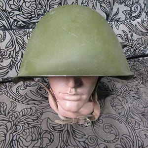 May include: An olive-green military helmet with a leather chin strap. The helmet is displayed on a mannequin head. The helmet has a rounded shape and a dark rim. The chin strap is tan and has a metal buckle.