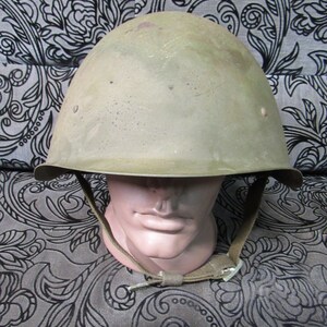 Puede incluir: Un casco militar verde oliva con ala y barboquejo. El casco se exhibe sobre una cabeza de maniquí. El casco tiene una apariencia ligeramente desgastada, lo que sugiere edad o uso. El barboquejo es de color beige claro.