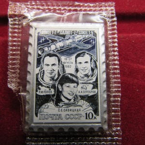 May include: A postage stamp featuring three cosmonauts and a spacecraft, with text in Cyrillic script. The stamp is encased in a clear plastic sleeve. The stamp commemorates the Soyuz T-7, Salyut 7, and Soyuz T-5 missions.