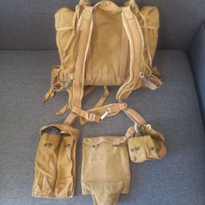 May include: A tan canvas backpack with multiple compartments and adjustable straps. Includes additional pouches and a waist belt. The backpack features metal buckles and a vintage aesthetic, suitable for outdoor activities or historical reenactments.