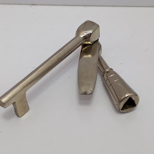 以下が含まれることがあります： 白い表面に配置された3つの銀色の金属工具。1つの工具はハンドルとT字型の端を持っています。もう1つは丸みを帯びたハンドルと平らな端を持っています。3つ目の工具は三角形の端を持っています。