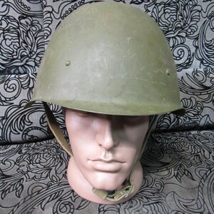 Puede incluir: Un casco militar verde oliva con barboquejo se muestra sobre una cabeza de maniquí. El casco tiene forma redondeada y una superficie ligeramente texturizada. El barboquejo es de color beige claro.