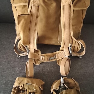 May include: A tan canvas backpack featuring adjustable shoulder straps, a waist belt, and two detachable pouches hanging from the bottom. One pouch contains a metal hand grenade with a pull ring, while the other pouch is empty and rectangular.