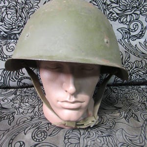 Puede incluir: Un casco militar verde oliva con una parte superior redondeada y un borde. El casco tiene una correa para la barbilla y se muestra en una cabeza de maniquí. La superficie del casco muestra signos de desgaste.
