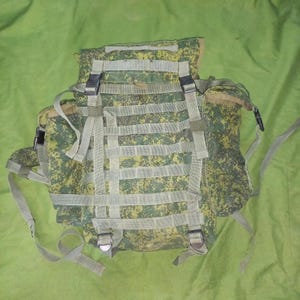 May include: A camouflage-patterned backpack with multiple compartments and straps. The backpack features a green, yellow, and brown digital camouflage design. It has multiple straps and buckles for securing items and adjusting fit. The bag is designed for outdoor activities.