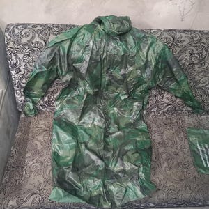 Op de afbeelding: Een groene camouflage regenjas met capuchon, uitgespreid op een patroon oppervlak. De regenjas is gemaakt van een glanzend, waterdicht materiaal. De jas heeft lange mouwen en een ontwerp over de hele lengte.