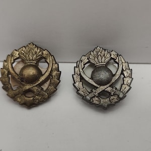 May include: Two metal badges with a thistle design. One badge is gold-toned, the other is silver-toned. Each badge features a central thistle, surrounded by a wreath and crossed swords.