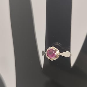 May include: A silver ring featuring a round, faceted, deep pink gemstone set in a scalloped, petal-like metal mounting. The band is smooth and tapers towards the setting, showcasing a polished metallic finish and a vibrant, central stone.