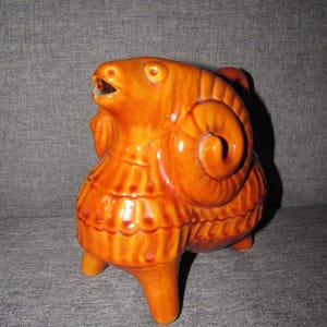May include: A ceramic ram figurine in a glossy orange glaze. The ram has detailed horns, a textured body, and is posed with its mouth open. The figurine has three legs and is a decorative item.